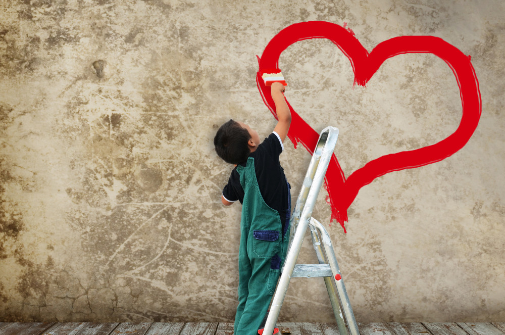 Kind malt ein Herz mit roter Farbe an eine Hauswand.