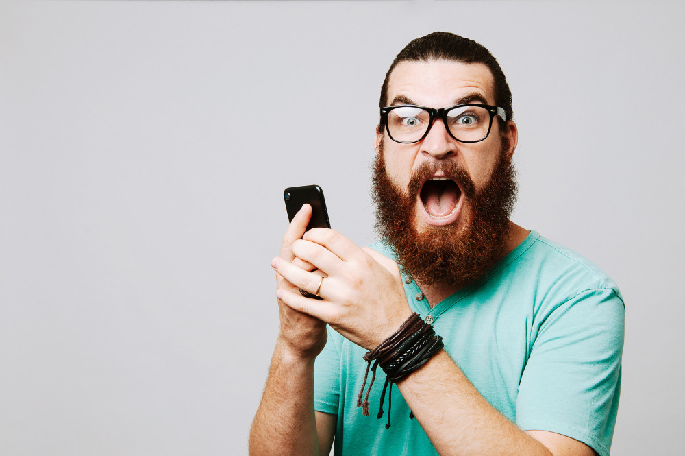 Junger Mann mit Brille schreit und hält ein Smartphone in der Hand Junger Mann mit Brille schreit und hält ein Smartphone in der Hand