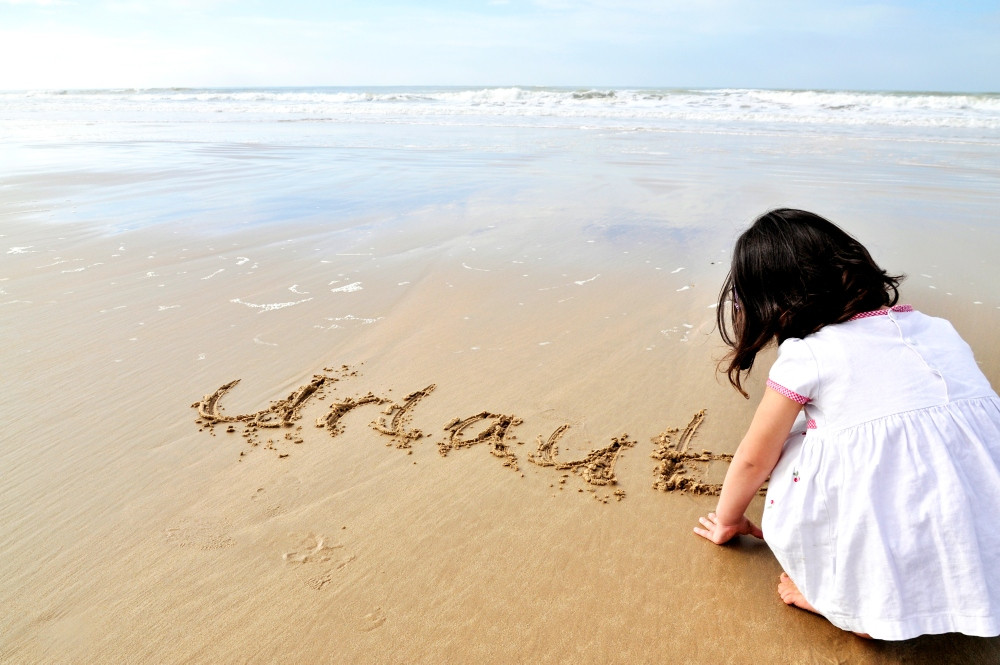 kleines Mädchen hockt am Strand und schreibt das Wort Urlaub in den Sand
