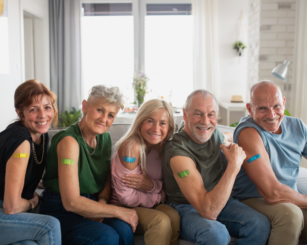 Gruppe von Senioren im Wohnzimmer auf der Couch mit bunten Pflastern an Einstichstelle am rechten Arm.