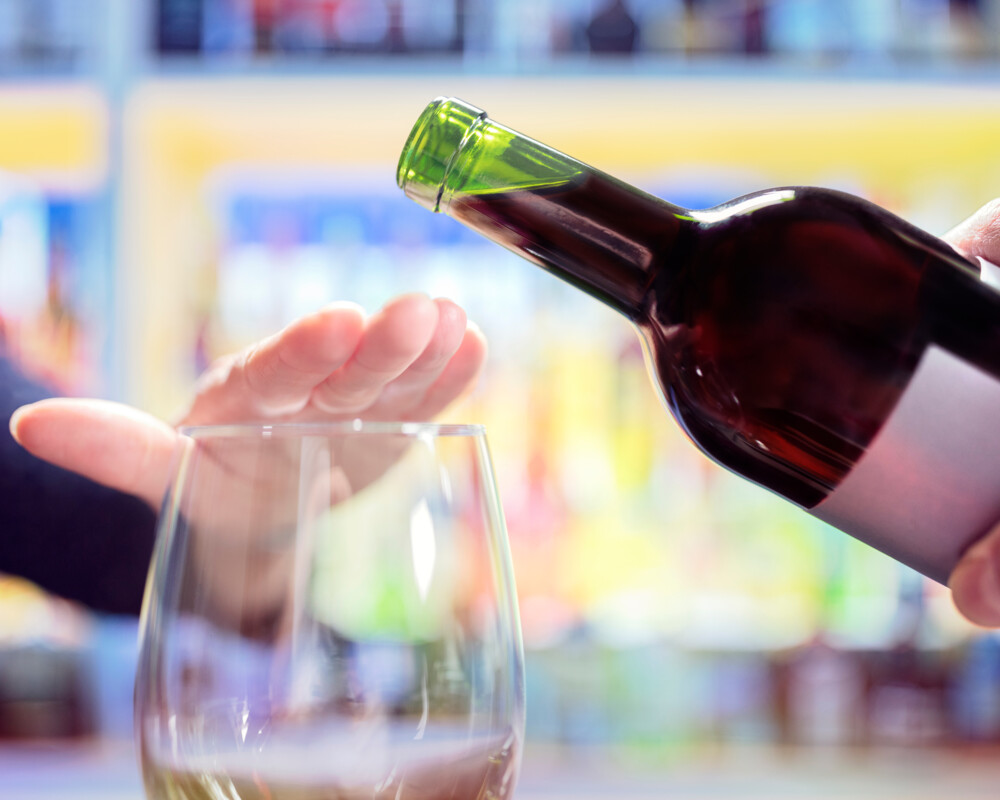 Weinflasche aus der in ein Glas Wein gegossen werden soll. Eine Hand legt sich abwehrend über das Glas.