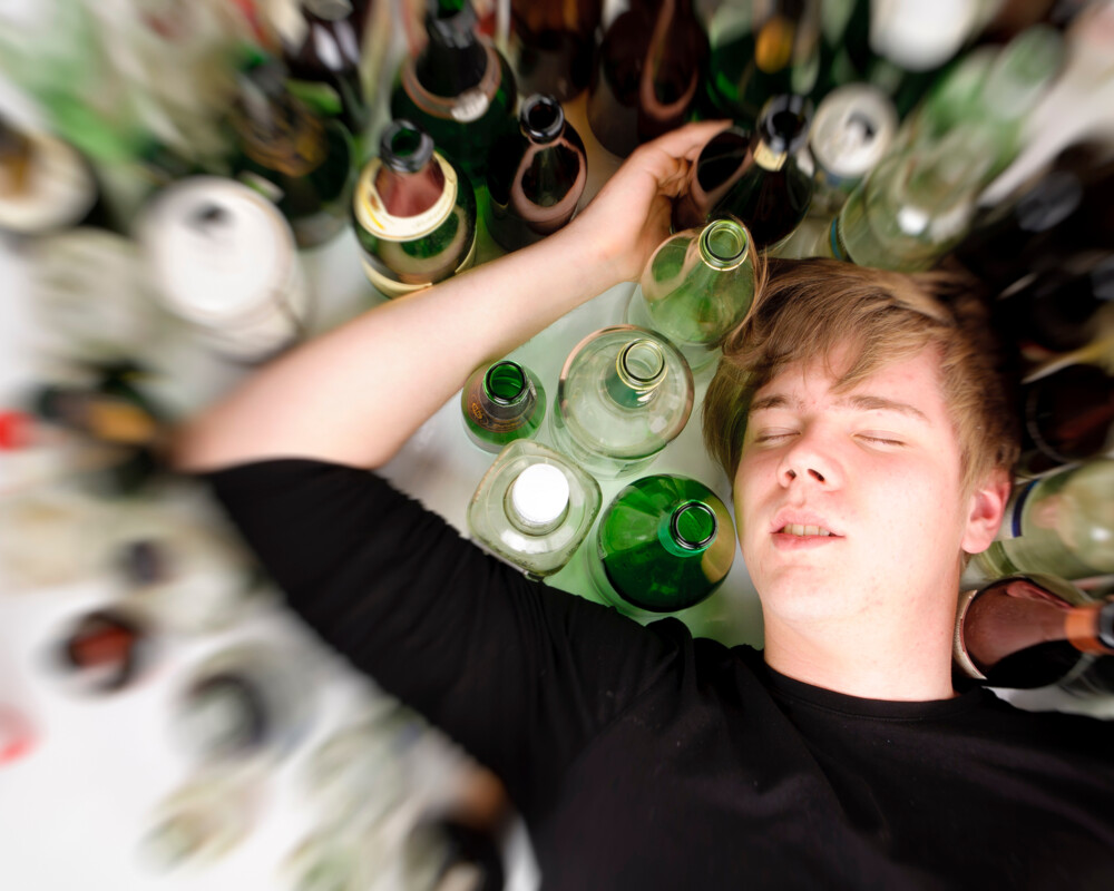 Junger Mann liegt mit geschlossenen Augenzwischen vielen Alkoholflaschen (Symbolbild)