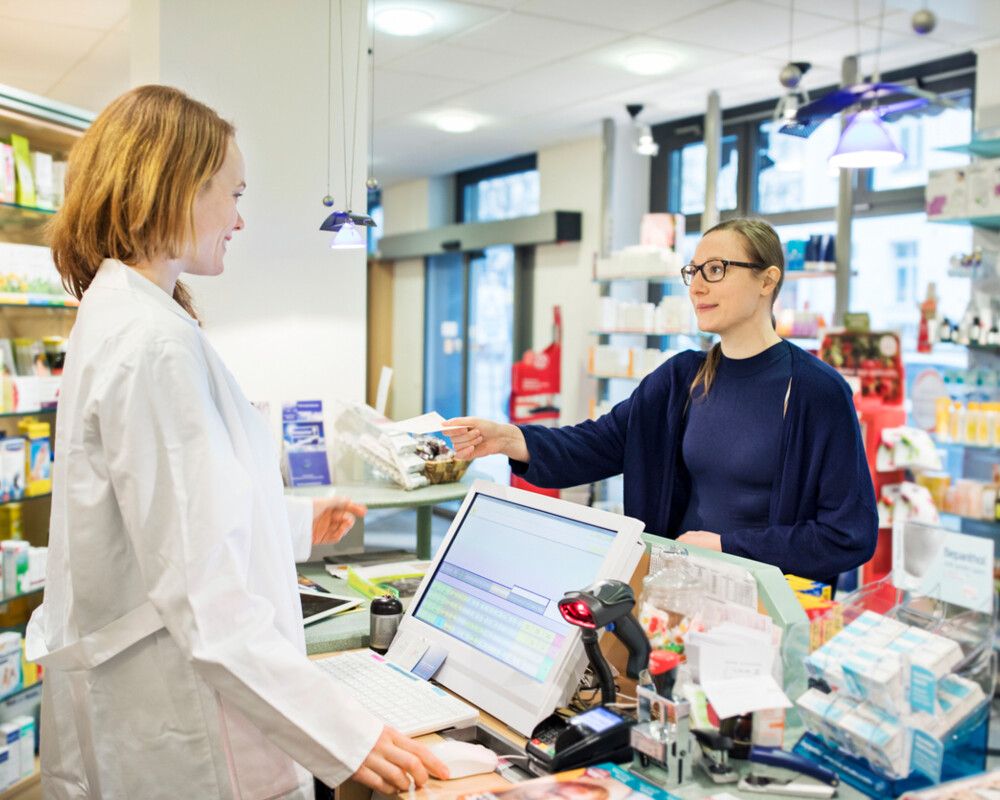 Kundin in der Apotheke reicht der PTA hinter dem HV-Tisch ein Rezept.