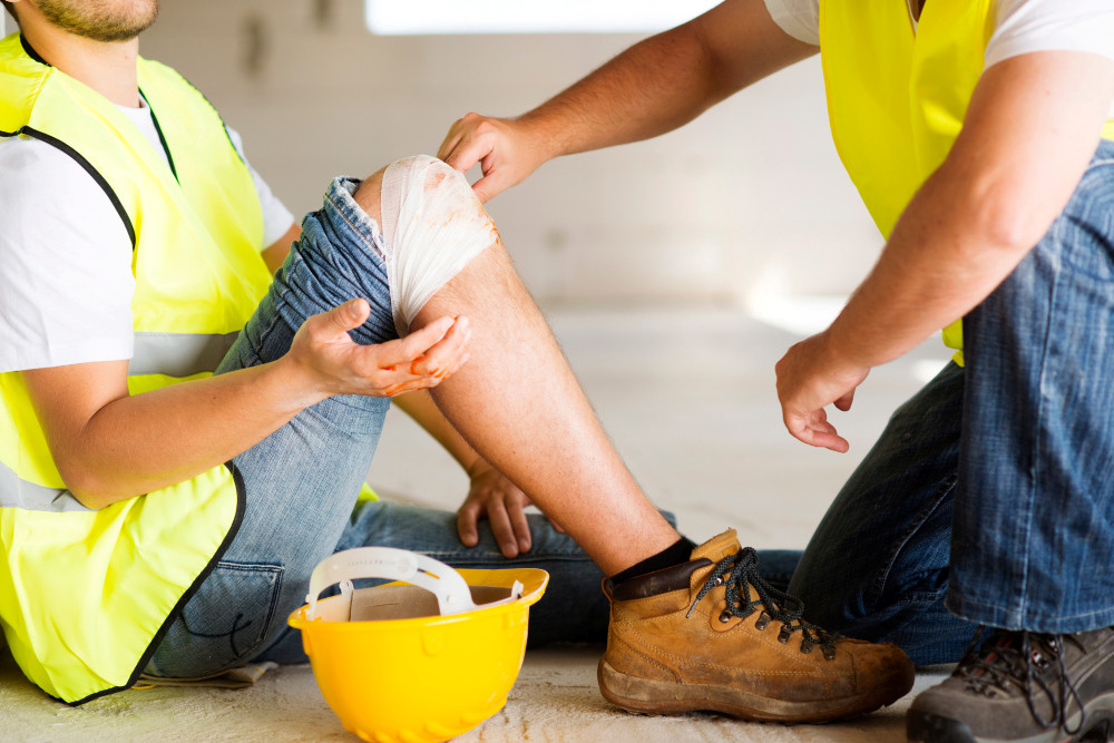 Verletzter Bauarbeiter mit verbundenem Knie wird von einem Kollegen betreut 