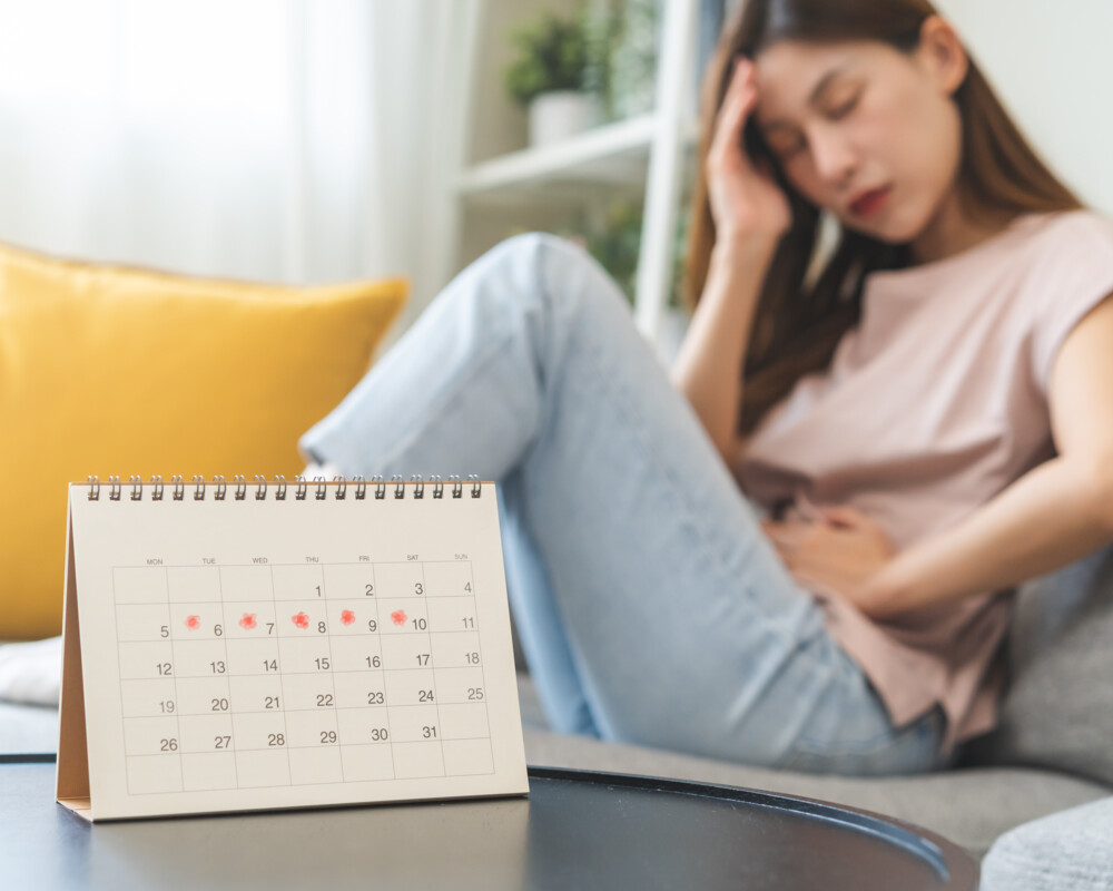 Junge Frau mit Bauch- und Kopfschmerzen auf dem Sofa im Hintergrund. Im Vordergrund ein Kalender mit fünf rot markierten Tagen. Junge Frau mit Bauch- und Kopfschmerzen auf dem Sofa im Hintergrund. Im Vordergrund ein Kalender mit fünf rot markierten Tagen.