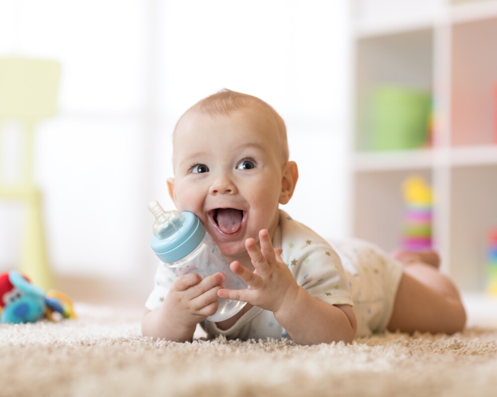 Lachendes Baby auf dem Bauch liegend mit einer Flasche in der Hand.