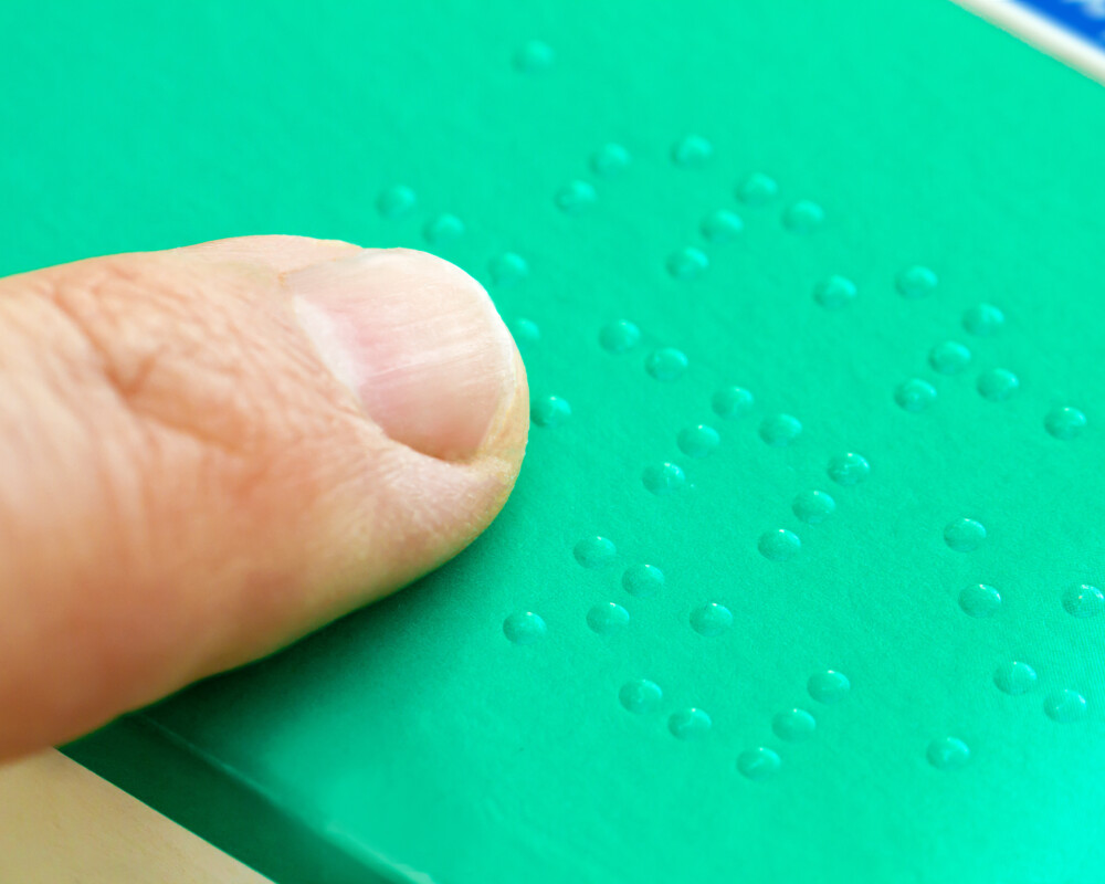Finger fährt über Brailleschrift auf Medikamentenpackung (grün), Symbolbild