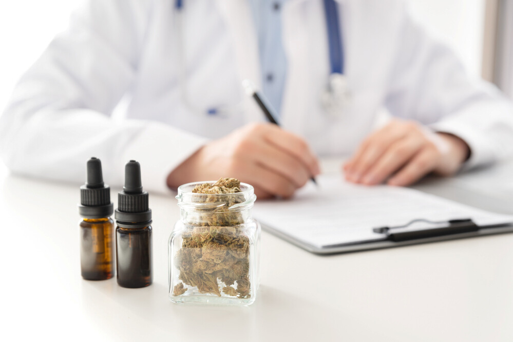 Cannabisblüten im Glas, Hanföl in Pipettenfläschchen und Arzt mit Stift in der Hand verschwommen im Hintergrund