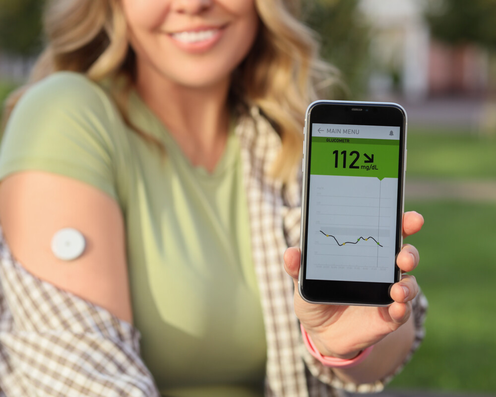 Frau mit CGM am rechten Oberarm zeigt Handy mit Wert 112 mg/dl in die Kamera