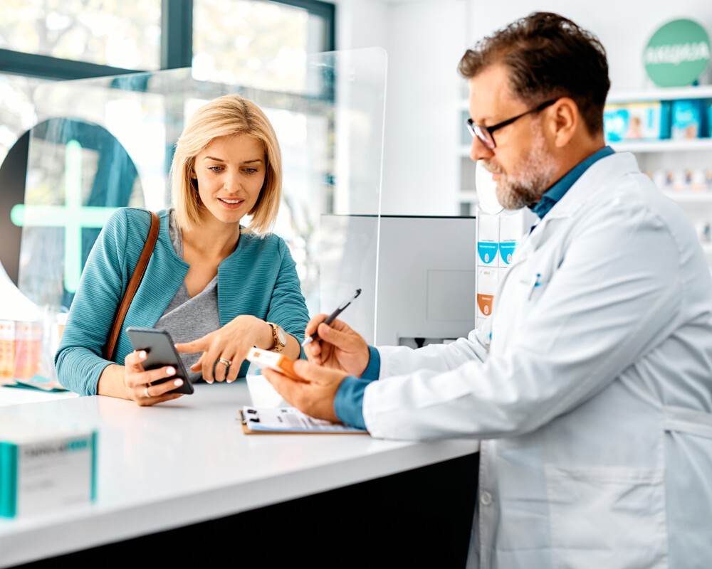 Eine Frau mittleren Alters, die ihr Handy benutzt, während sie sich mit einem Apotheker / PTA unterhält und in der Apotheke Medikamente kauft.