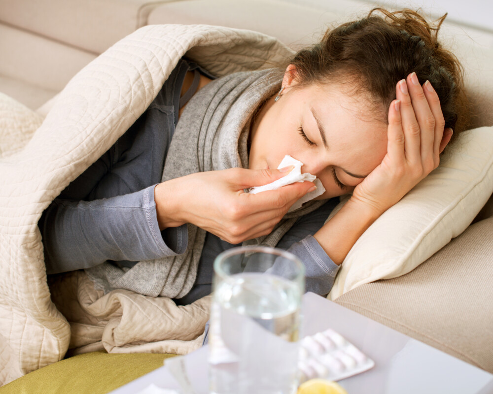 
Kranke Frau liegt auf dem Sofa und putzt sich die Nase. Im Vordergrund ein Wasserglas und ein Tablettenblister.