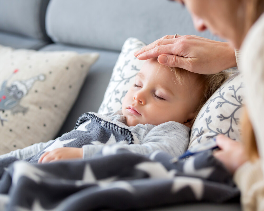 Mutter fühlt die Stirn bei ihrem kleinen Kinde, das mit geschlossenen Augen und zugedeckt auf dem Sofa liegt.