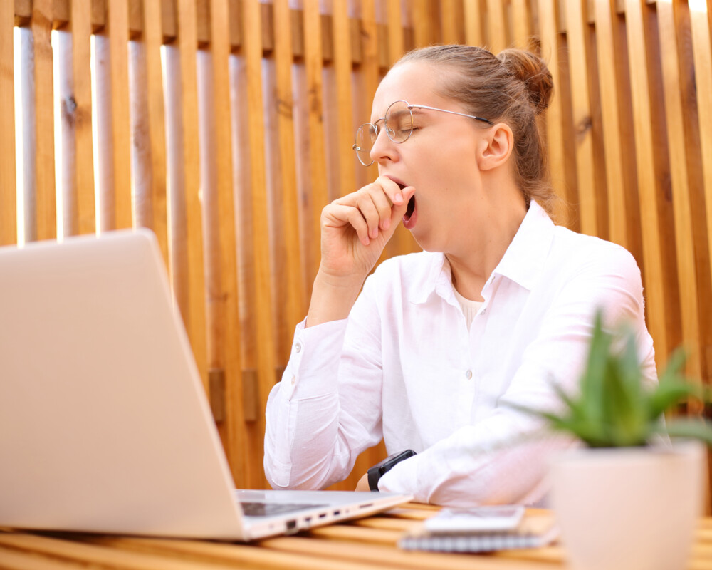 Junge Frau sitzt auf der Terrasse mit Laptop und gähnt.