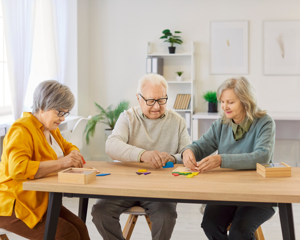 Zwei ältere Frauen und ein älterer Mann sitzen am Tisch und legen ein einfaches Puzzle zusammen.