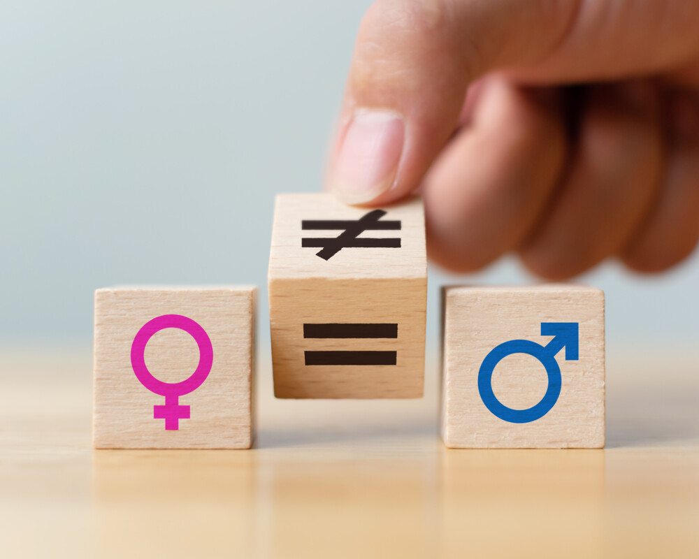 Drei Holzwürfel. Linker zeigt pinkes Zeichen für Frauen, der rechte ein blaues Zeichen für Männern. In der Mitte gekippter Würfel mit Gleichheitszeichen und Ungleichheitszeichen.
