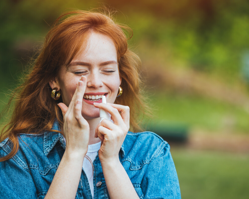 Rothaarige Frau wendet lächelnd ein Nasenspray gegen Heuschnupfen an.