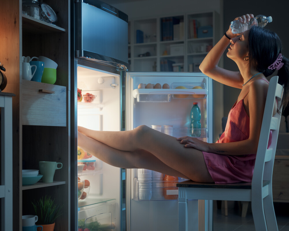 Frau sitzt im abgedunkelten Raum vor geöffnetem Kühlschrank, die Füße im Kühlschrank, ein Wasserflasche zur Kühlung an die Stirn gelegt.