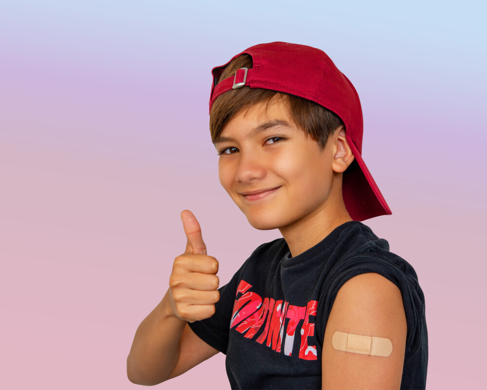 11- bis 12-jähriger Junge mit roter Baseballkappe, Pflaster auf dem linken Arm. Er lächelt in die Kamera und zeigt Daumen nach oben.