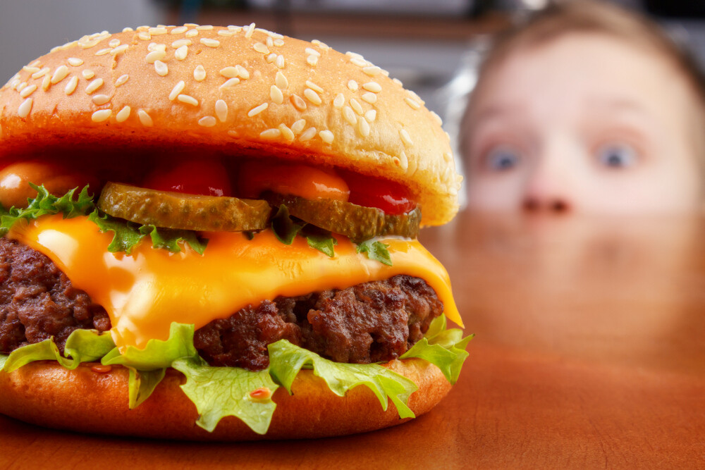 Hungriger Junge mit weit aufgerissenen Augen starrt auf einen Cheeseburger (scharf im Vordergrund) Hungriger Junge mit weit aufgerissenen Augen starrt auf einen Cheeseburger (scharf im Vordergrund)