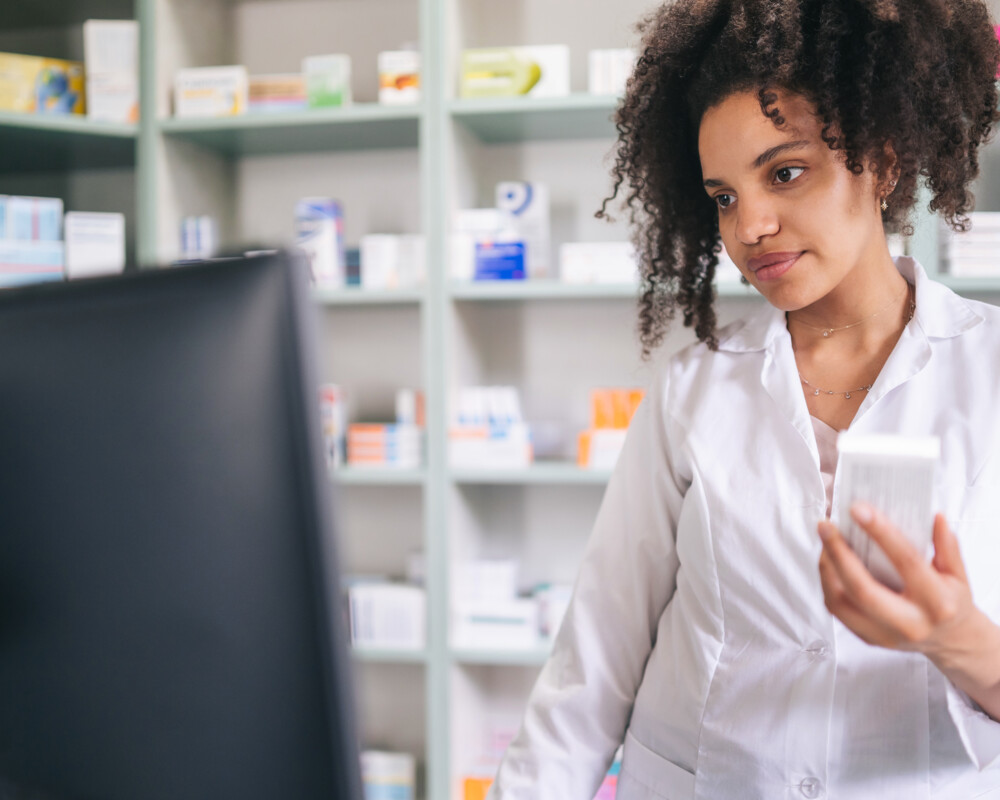 Apothekenangestelle mit einer Packung in der Hand schaut im Computer etwas nach.