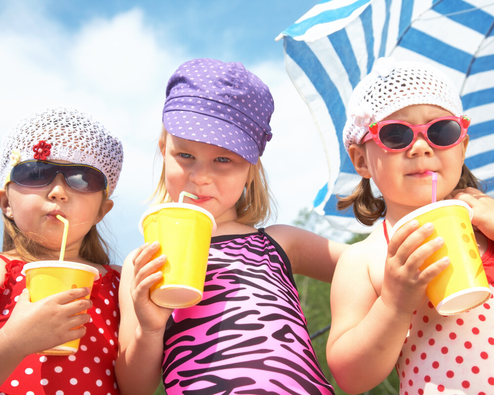 Drei Kinder mit Sonnenkappen, zwei mit Sonnenbrille, alle halten Getränk mit Strohhalm in der Hand