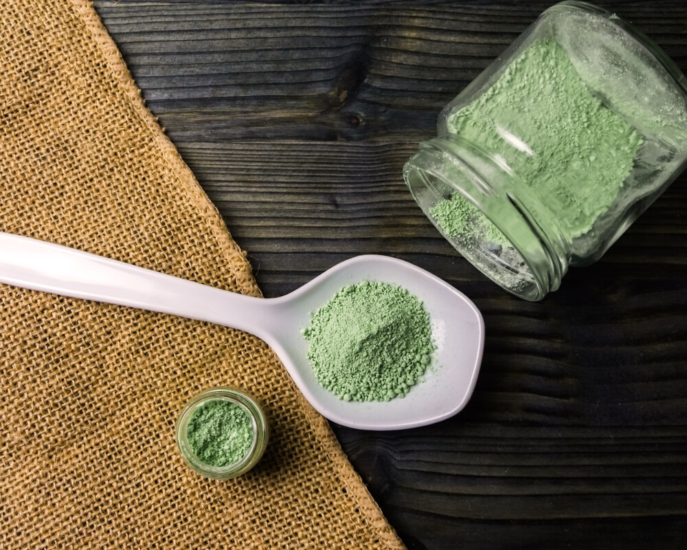 Kratompulver auf einem weißen Keramiklöffel und in einem umgekippten Glas Kratompulver auf einem weißen Keramiklöffel und in einem umgekippten Glas