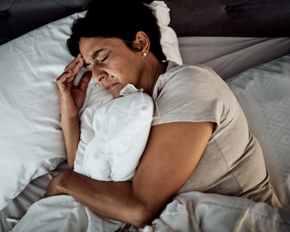 Frau mit Hand am Kopf liegt im Bett auf einem weißen Kopfkissen im abgedunkelten Raum.