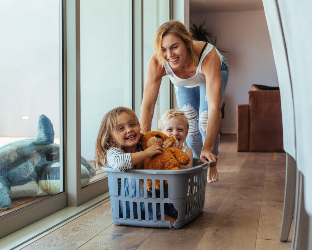 Mutter schiebt Sohn und Tochter in einem Wäschekorb durchs Zimmer.