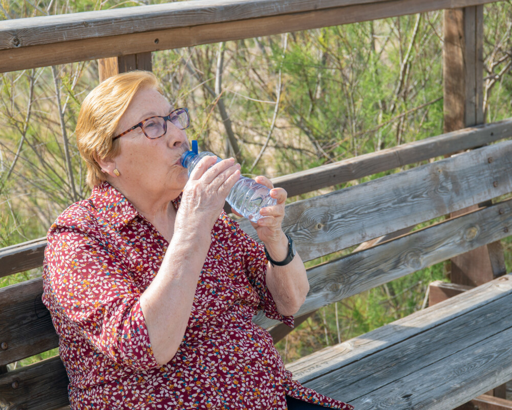 Seniorin sitzt auf einer Bank und trinkt aus einer Wasserflasche.