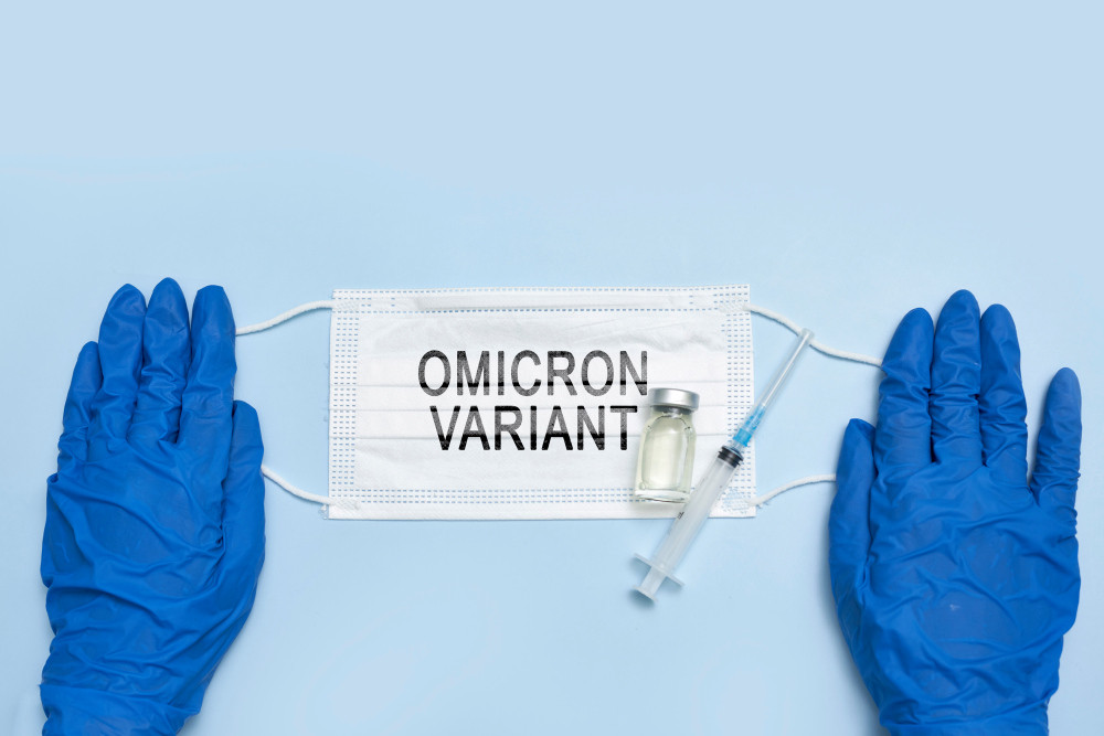 OP-Maske mit Aufdruck Omicron Variant sowie Vial und Spritze wird von zwei blau behandschuhten Händen gehalten