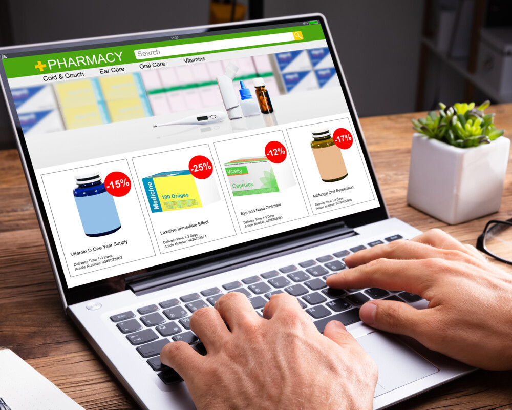 Hände auf der Tastatur eines geöffneten Laptops. Bildschirm zeigt symbolisch die Seite einer Online-Apotheke.