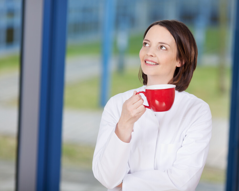 Junge Frau mit roter Tasse lehnt lächelnd am Fenster.