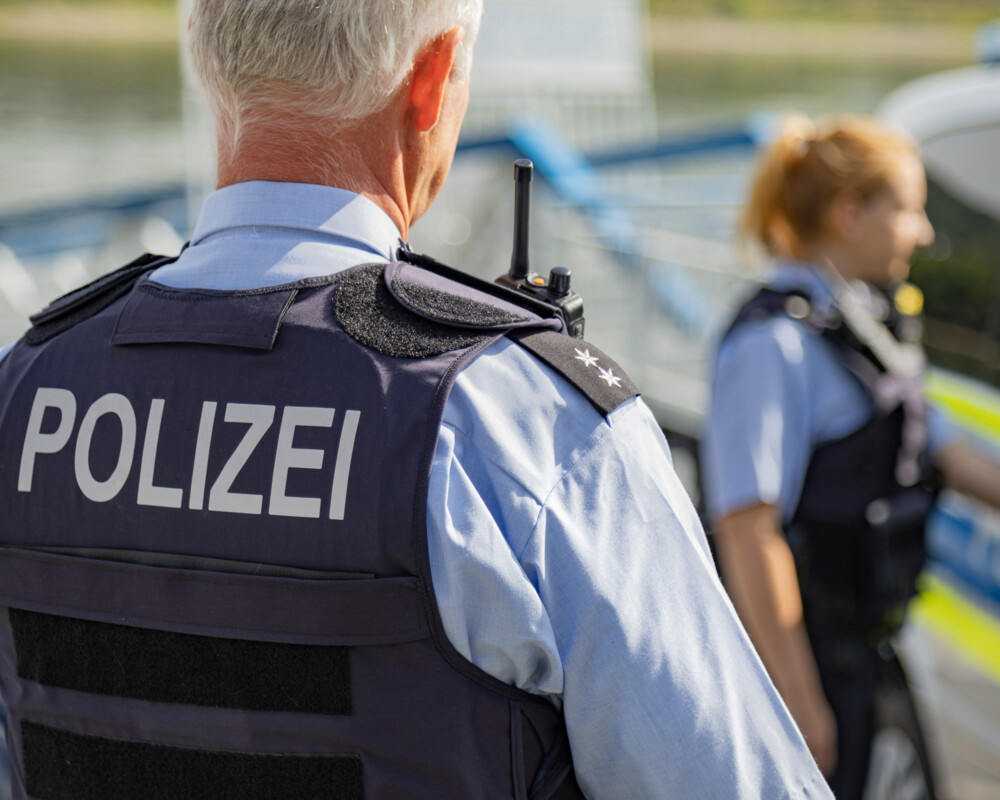 Älterer männlicher Polizist von hinten mit Aufschrift „Polizei“ auf dem Rücken. Im Hintergrund verschwommen eine Polizistin mit Pferdeschwanz.