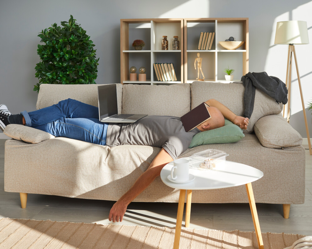 Mann schläft auf einem Sofa, mit Laptop auf dem Bauch und Buch über dem Gesicht.