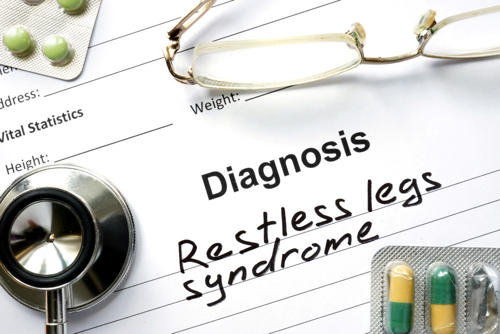 Zettel mit Diagnos „Restless less syndrome“, daraufliegend eine Lesebrille, ein Stethoskop und zwei Blister mit Tabletten und Kapseln