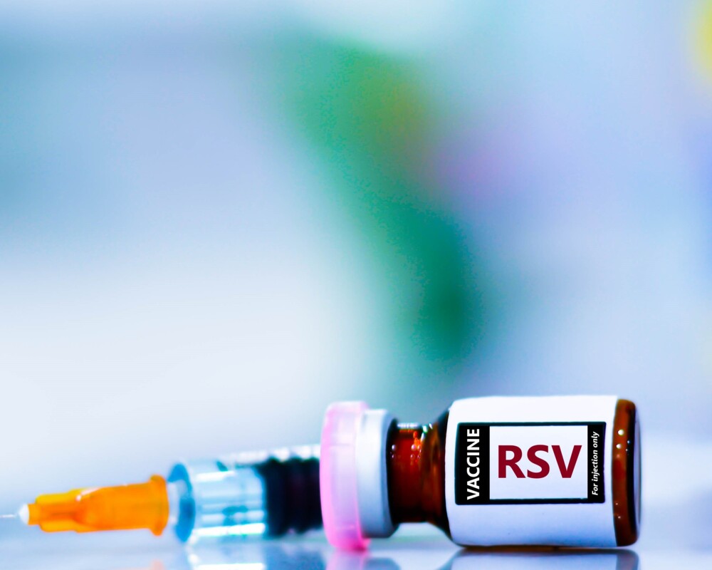 Liegendes Vial mit Aufschrift „Vaccine RSV“ und im Hintergrund eine Spritze Liegendes Vial mit Aufschrift „Vaccine RSV“ und im Hintergrund eine Spritze