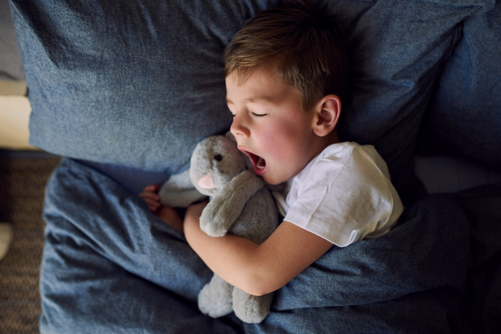 Kleiner Junge mit Kuschelhasen im Arm liegt mit geschlossenen Augen im Bett und gähnt