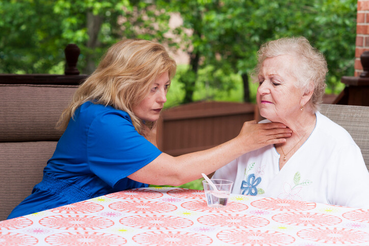Pflegekraft hilft Seniorin beim Schlucken durch das Drücken mit der Hand auf den Kehlkopf, beide sitzen an einem Tisch auf dem Balkon.