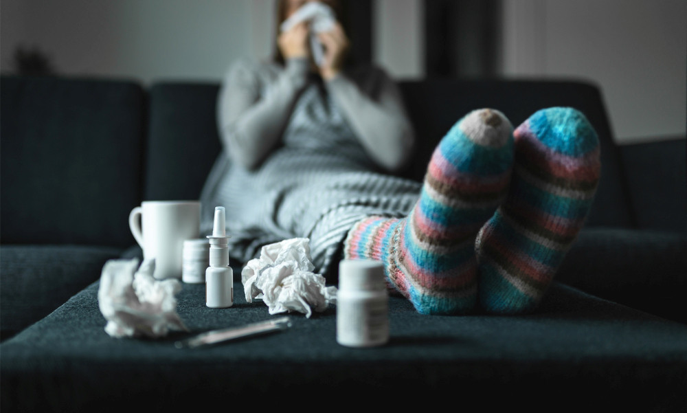 Frau auf dem Sofa, im Vordergrund genutzte Taschentücher, Nasenspray, Tablettendose und Teebecher