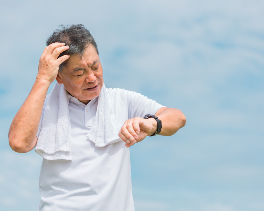 Asiatischer älterer Mann mit weißem T-Shirt und Handtuch um den Hals schaut auf seinen Fitness-Tracker.