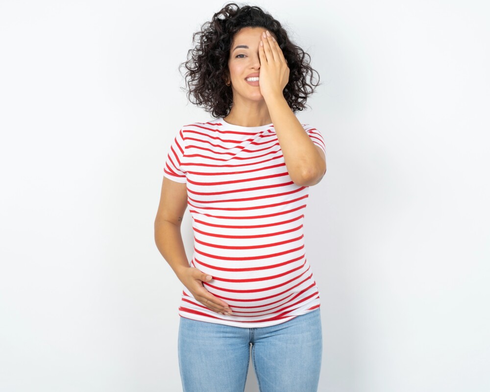 Schwangere Frau in einem weiß-rot-gestreiften T-Shirt hält sich mit der linken Hand das linke Auge zu.