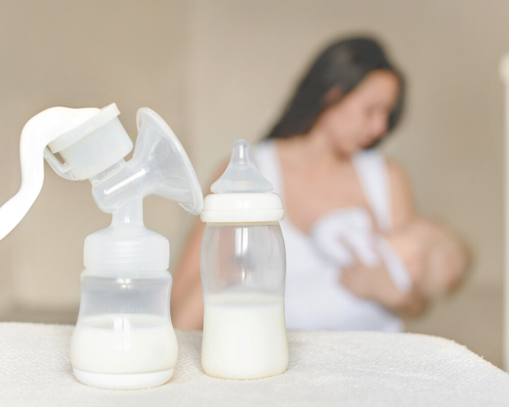 Frau stillt ihr Baby (unscharf im Hintergrund). Im Vordergrund zwei Fläschchen mit Milch gefüllt, eines mit Aufsatz einer Hand-Milchpumpe.