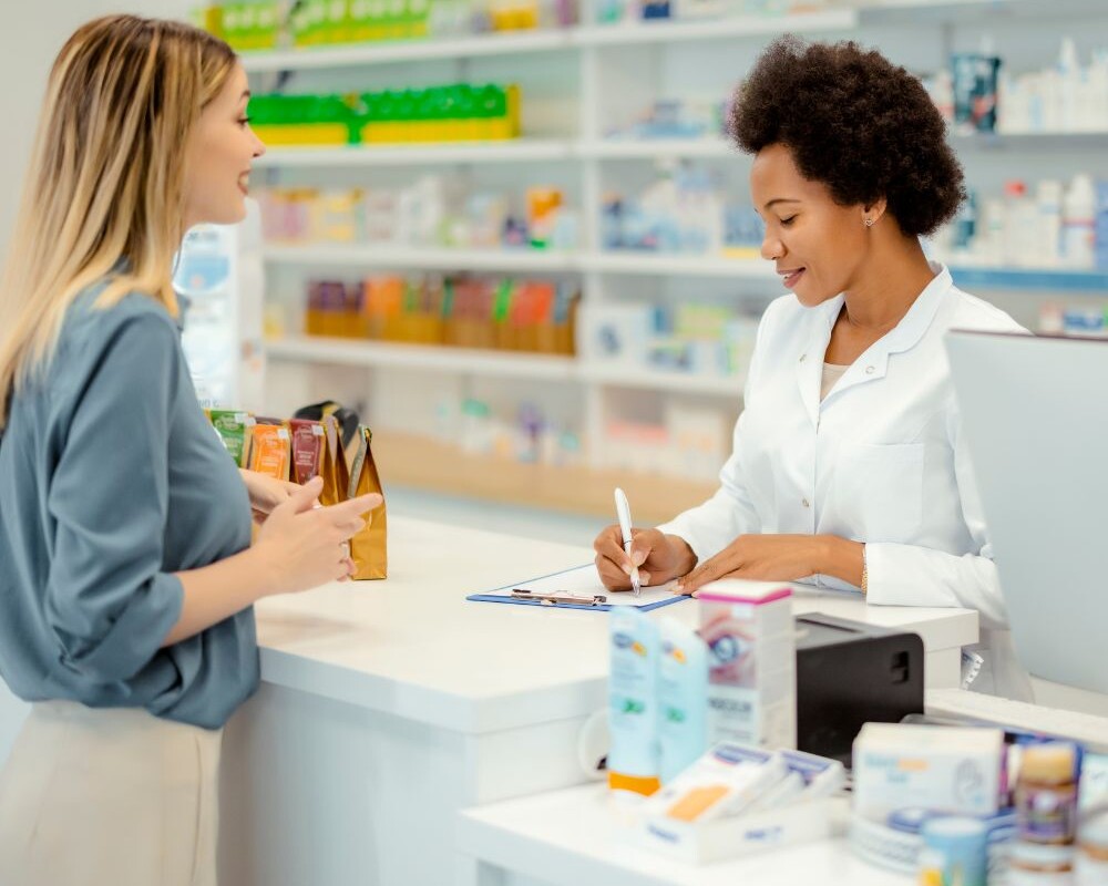 Zwei weibliche Personen in einer Apotheke: Eine steht hinter dem HV-Tisch in einem weißen Kittel und notiert gerade etwas, die andere steht mit hellem Rock und dunkelgrüner Bluse bekleidet davor.