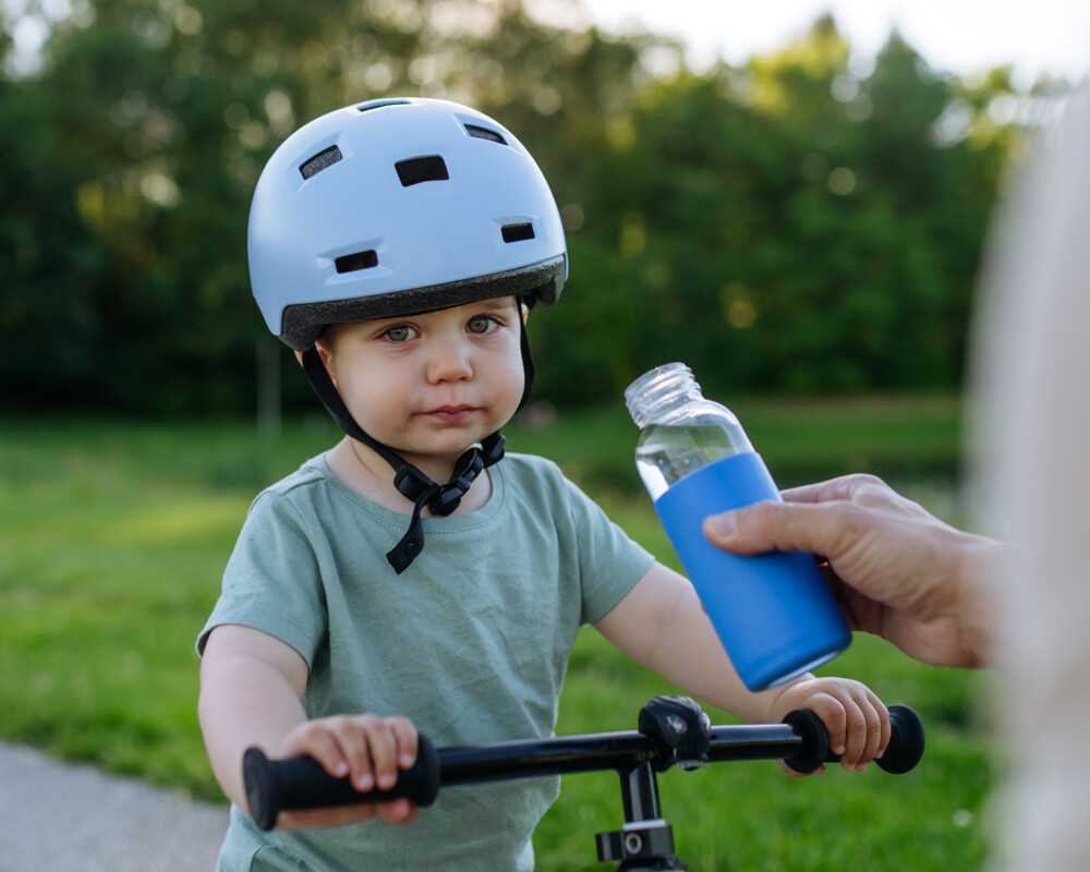 Kleiner Junge mit blauem Fahrradhelm blickt skeptisch in die Kamera. Eine Hand hält ihm eine Flasche mit Wasser hin.