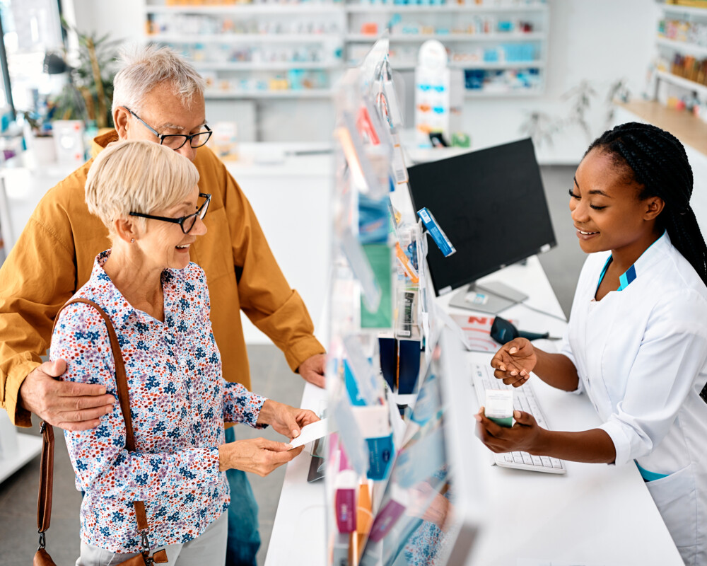 Seniorenpaar holt Medikamente in der Apotheke, PTA berät hinter dem HV-Tisch.