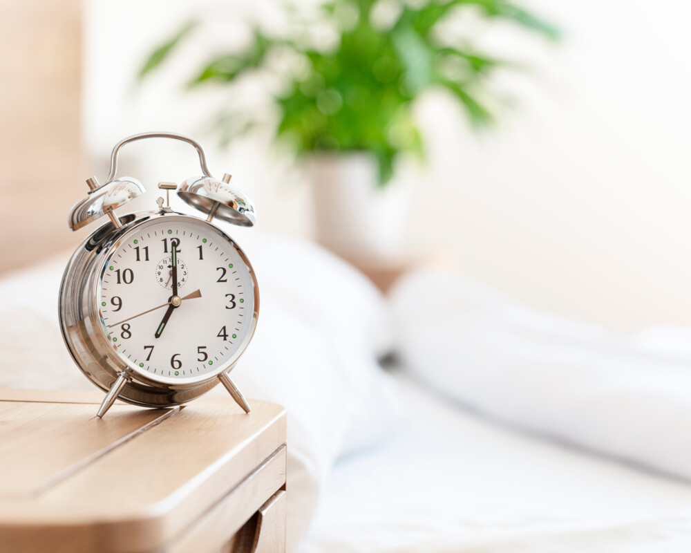 Retro-Wecker auf dem Nachttisch zeigt 7:00 Uhr an, im Hintergrund ein Bett.