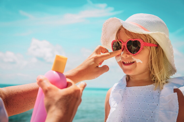 Kind mit Sonnenbrille und Sonnenhut bekommt Sonnencreme im Gesicht aufgetragen. Kind mit Sonnenbrille und Sonnenhut bekommt Sonnencreme im Gesicht aufgetragen.