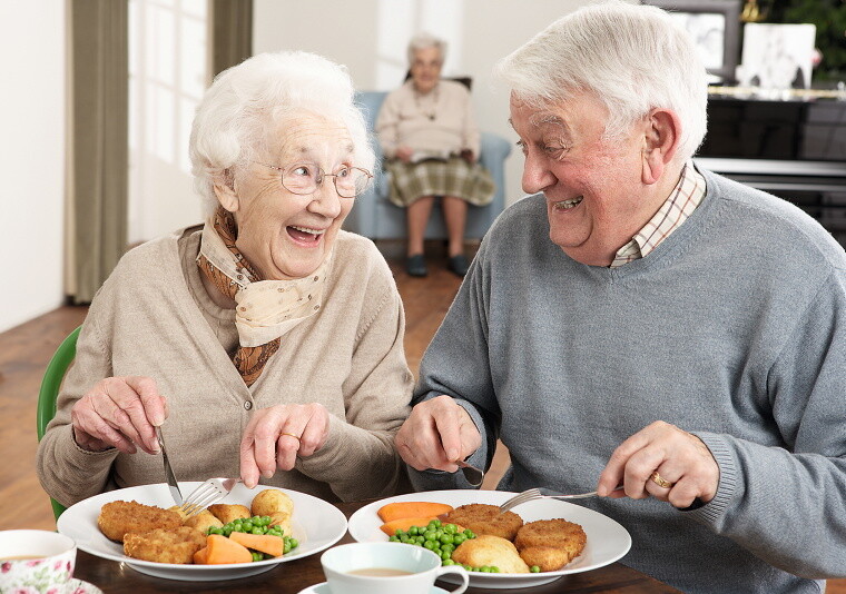Ältere Frau (links) und älterer Mann (rechts) lachen beim Essen.