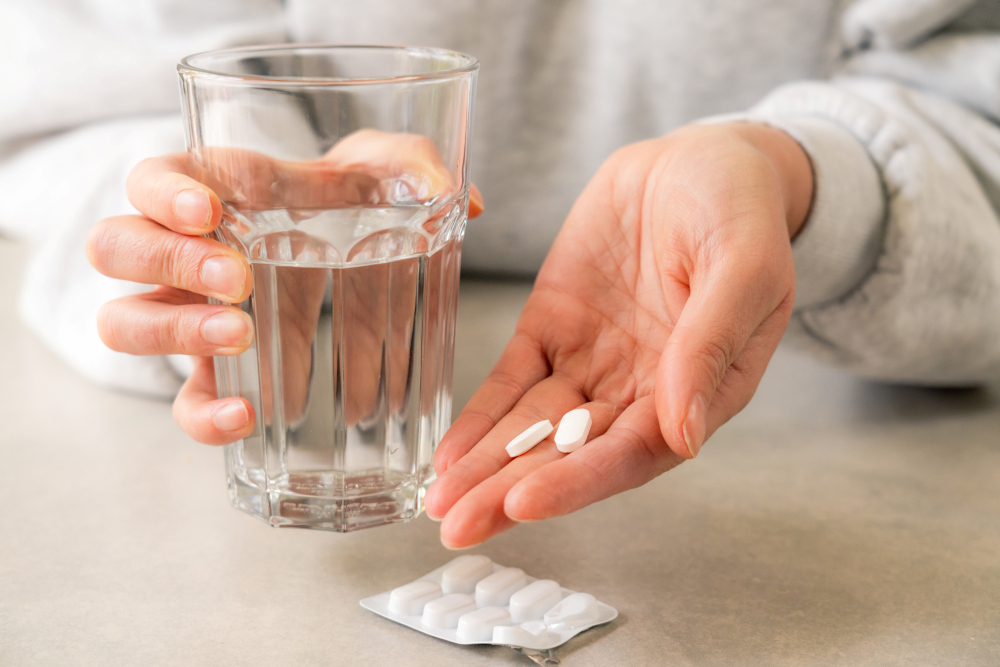 Frau hält Wasserglas in der einen Hand, in der anderen zwei Tabletten. Auf dem Tisch ein Blister mit Tabletten.