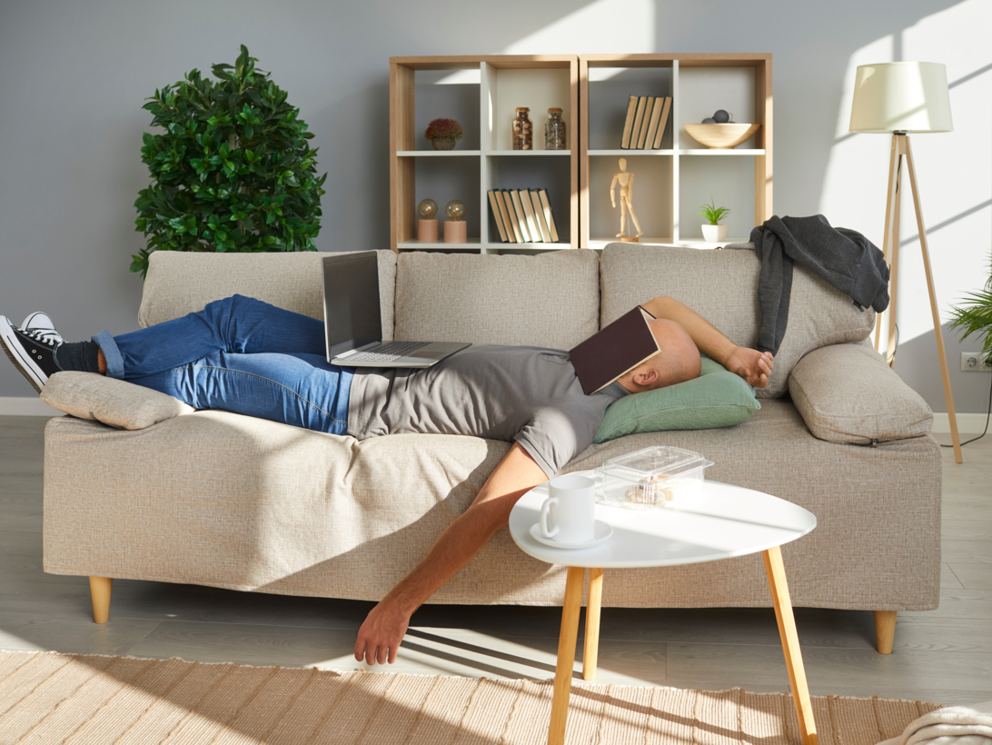 Mann schläft auf einem Sofa, mit Laptop auf dem Bauch und Buch über dem Gesicht.